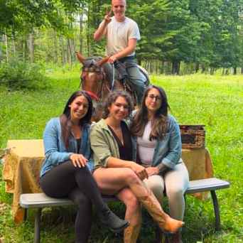 Three women sitting on a bench, man on horseback behind them in a grassy, wooded area.
