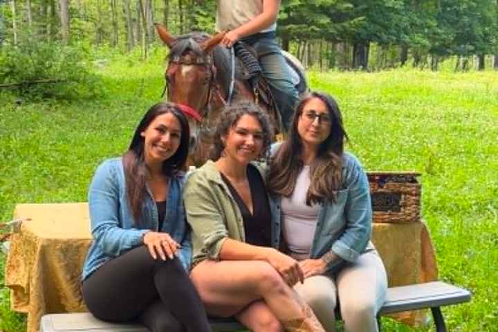 Three women sitting on a bench, man on horseback behind them in a grassy, wooded area.