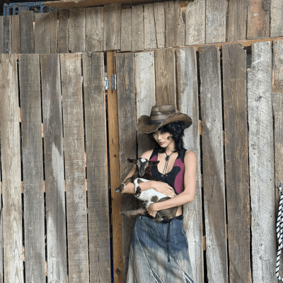 Person in a cowboy hat holding a small goat in front of a wooden fence.