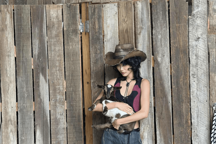 Person in a cowboy hat holding a small goat in front of a wooden fence.