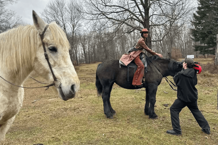 Person on black horse with red saddle, another holding reins, white horse in foreground, bare trees in background.