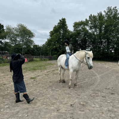 Person sitting on a white horse while another person takes a photo in an outdoor area with trees.