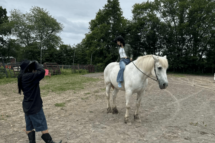 Person sitting on a white horse while another person takes a photo in an outdoor area with trees.