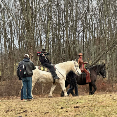 People filming horseback riders in a wooded area on a cloudy day.