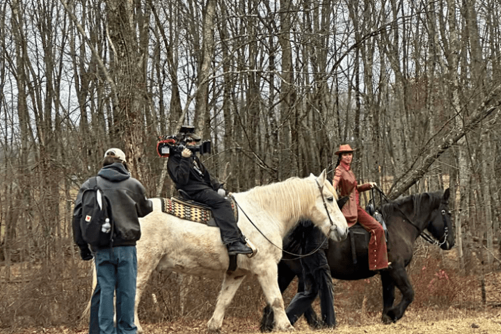 People filming horseback riders in a wooded area on a cloudy day.