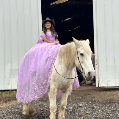 Girl in purple dress and cowboy hat on a white horse in front of barn.