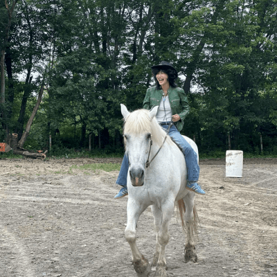 Person in cowboy hat riding a white horse on a dirt path with trees in the background.