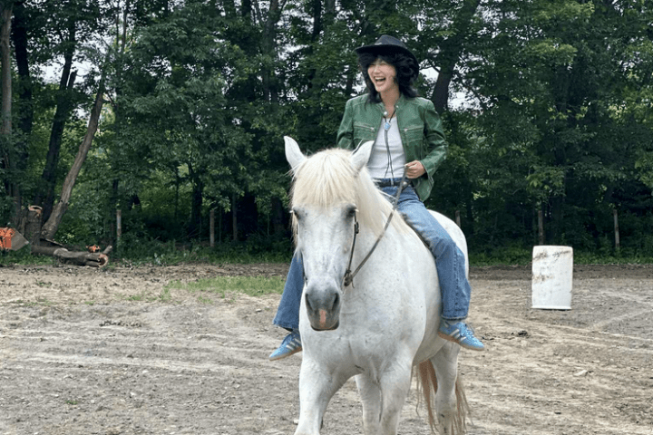 Person in cowboy hat riding a white horse on a dirt path with trees in the background.