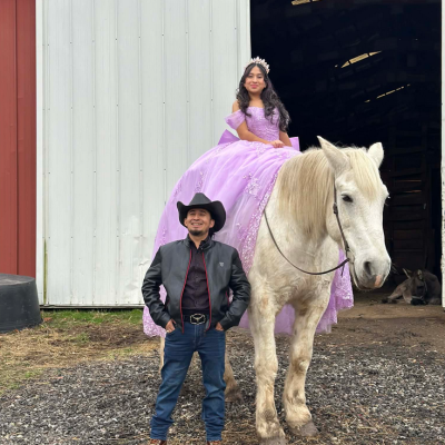 Girl in purple gown on horse, man in cowboy attire, barn background.