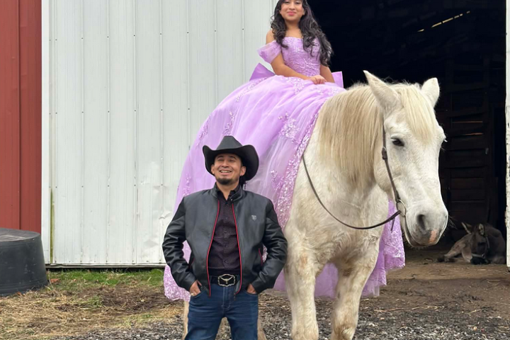 Girl in purple gown on horse, man in cowboy attire, barn background.