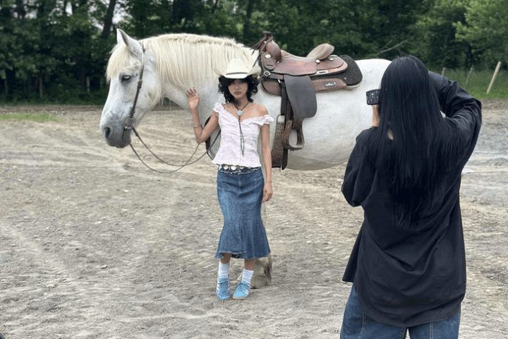 Person in cowboy hat poses with white horse while another person takes a photo in an outdoor setting.