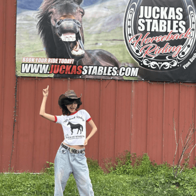 Person in cowboy hat pointing at horse stables sign on red barn wall.