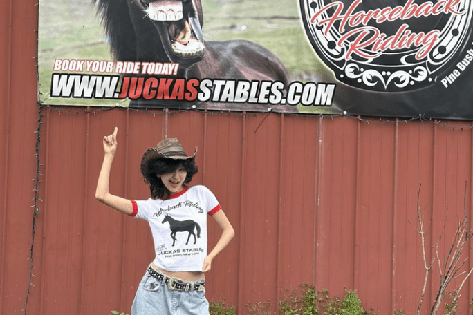 Person in cowboy hat pointing at horse stables sign on red barn wall.
