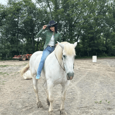 Person in cowboy hat riding a white horse in a forested area with a tractor in the background.