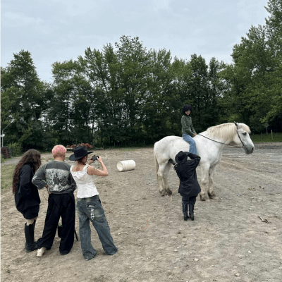 Person on white horse being photographed by four others in an outdoor setting with trees.