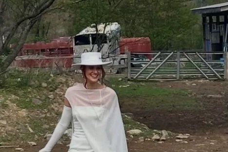 Woman in white dress and hat walking outdoors by farm.