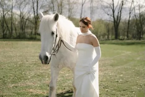 Bride in white dress walking with a white horse in a grassy field.