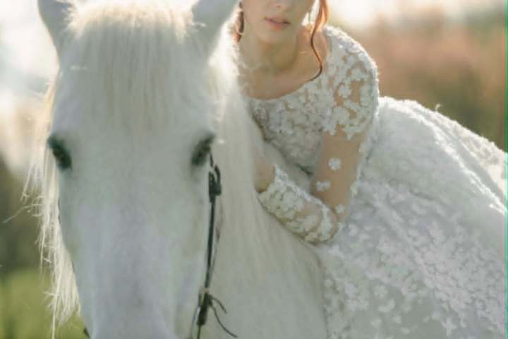 Woman in lace dress riding white horse in a grassy field.