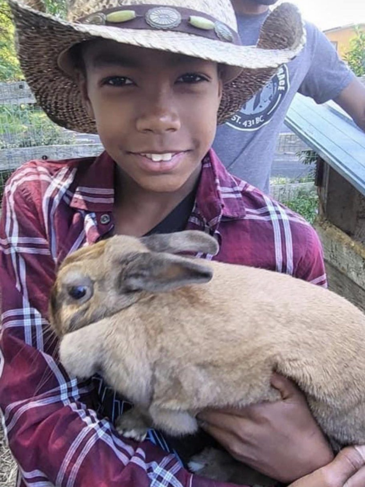 Child in a straw cowboy hat holds a rabbit, with a person in a star hat in the background.