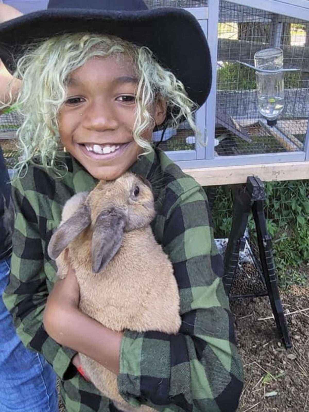 Child with curly hair and cowboy hat smiling while holding a brown rabbit outdoors.