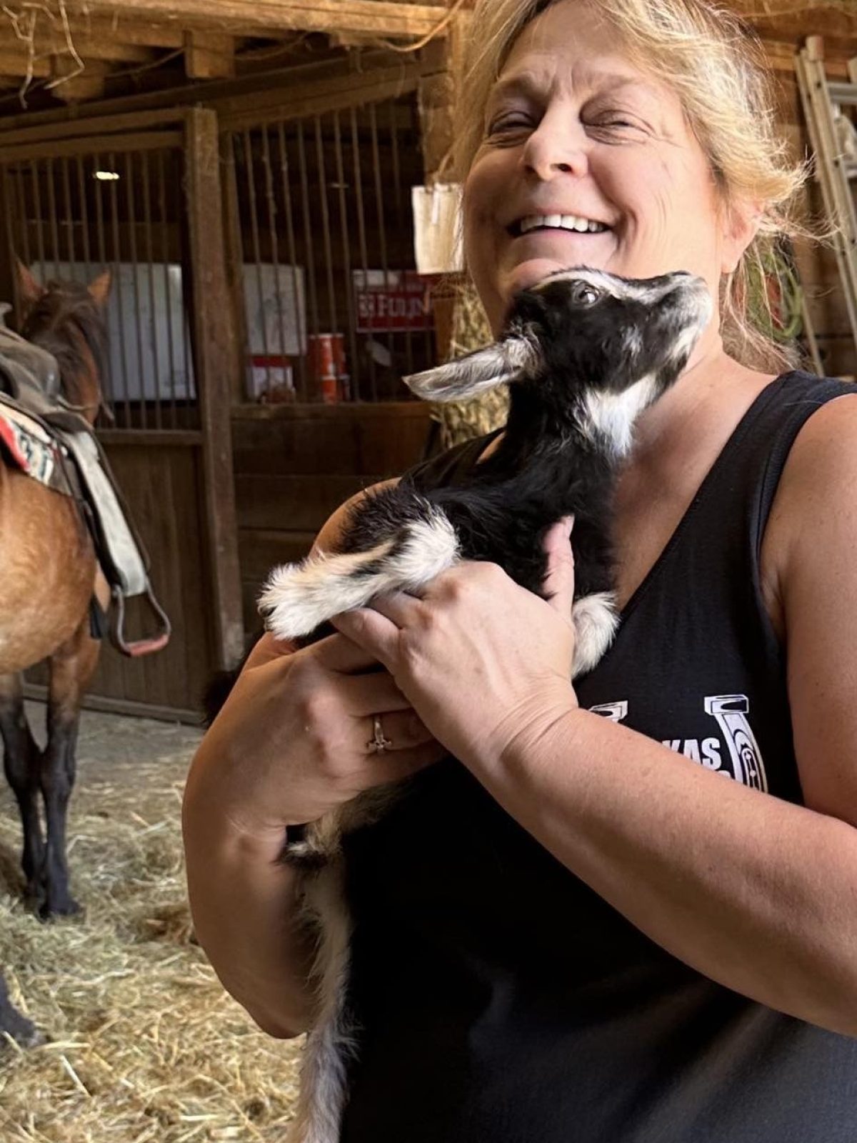 Woman happily holding a small goat in a barn with hay bales.