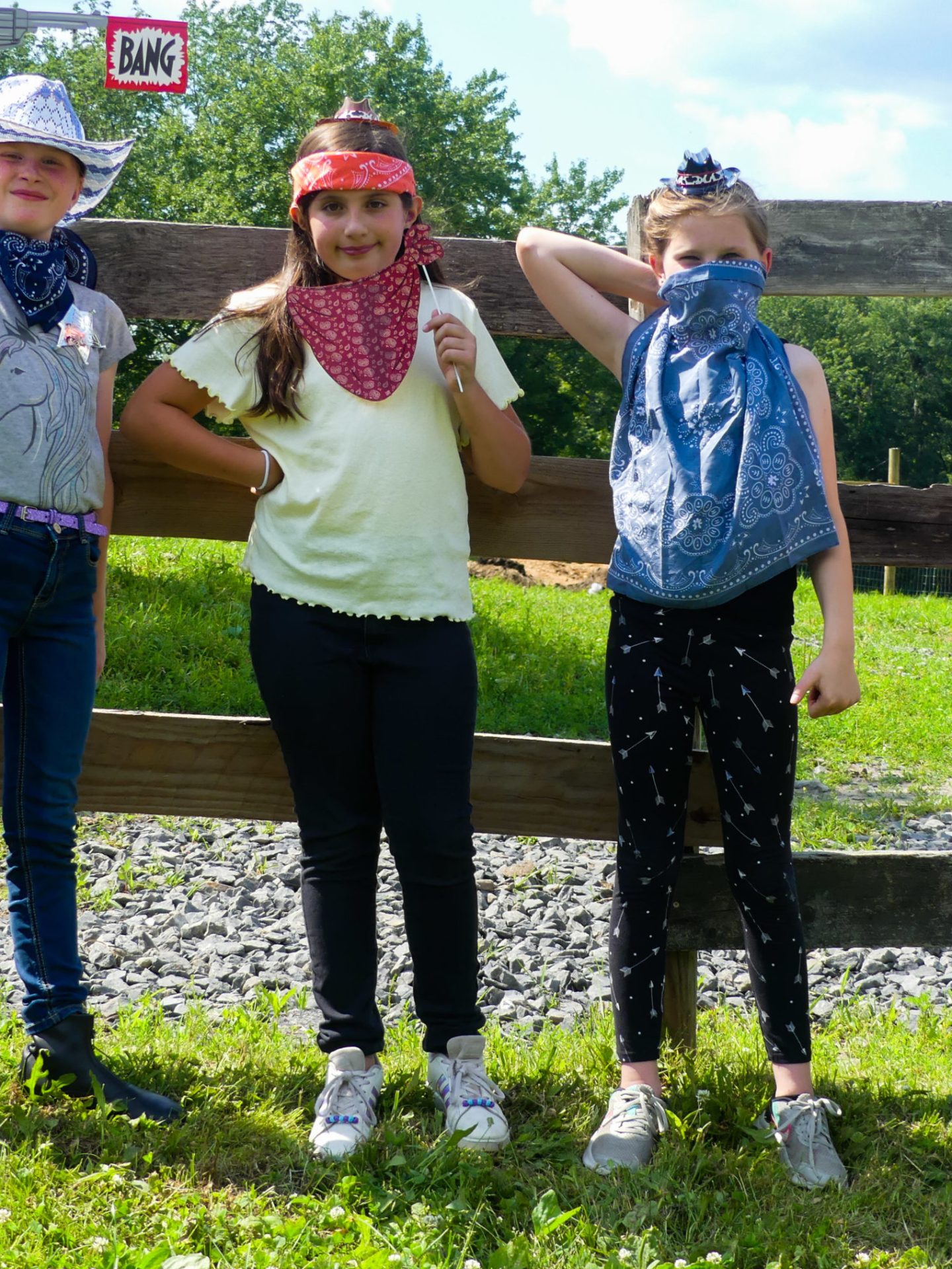 Four kids in cowboy attire pose with hats and bandanas outdoors.