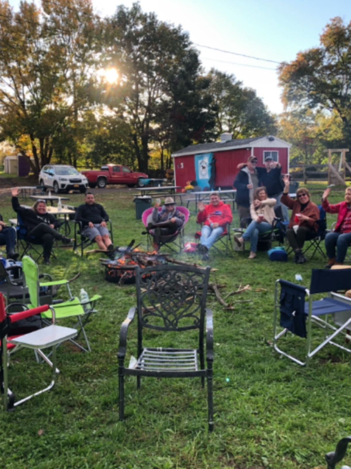Group of people sitting around a campfire with chairs on grass during a sunny day.
