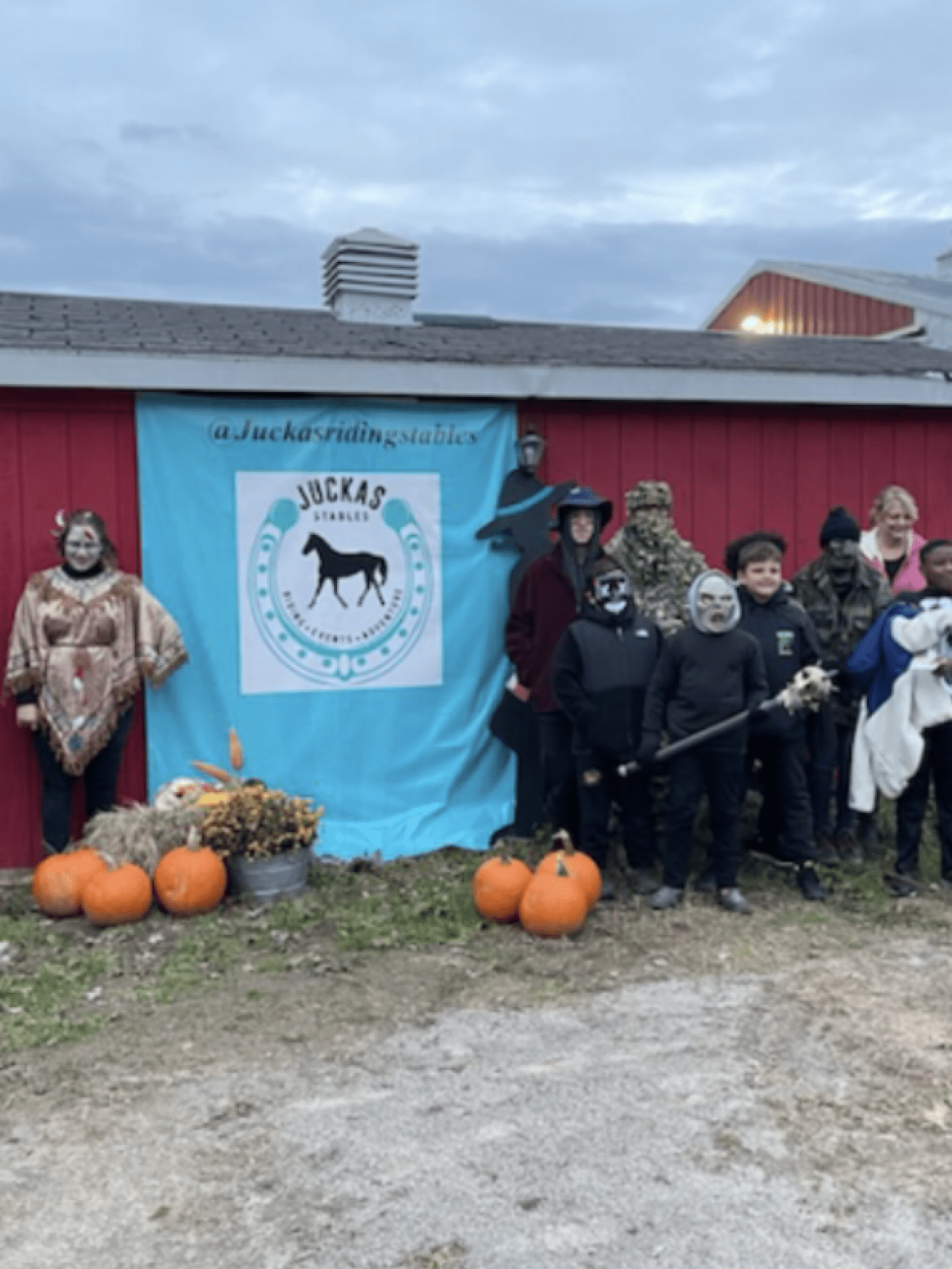 Group in costumes stands by a red barn with a blue 'Juckas Stables' banner and pumpkins.