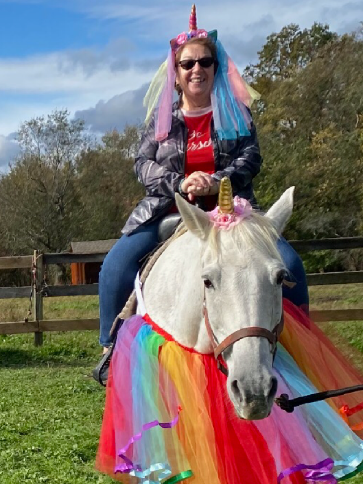 Person on a white horse with rainbow skirt and unicorn horn headbands on both, outdoors.