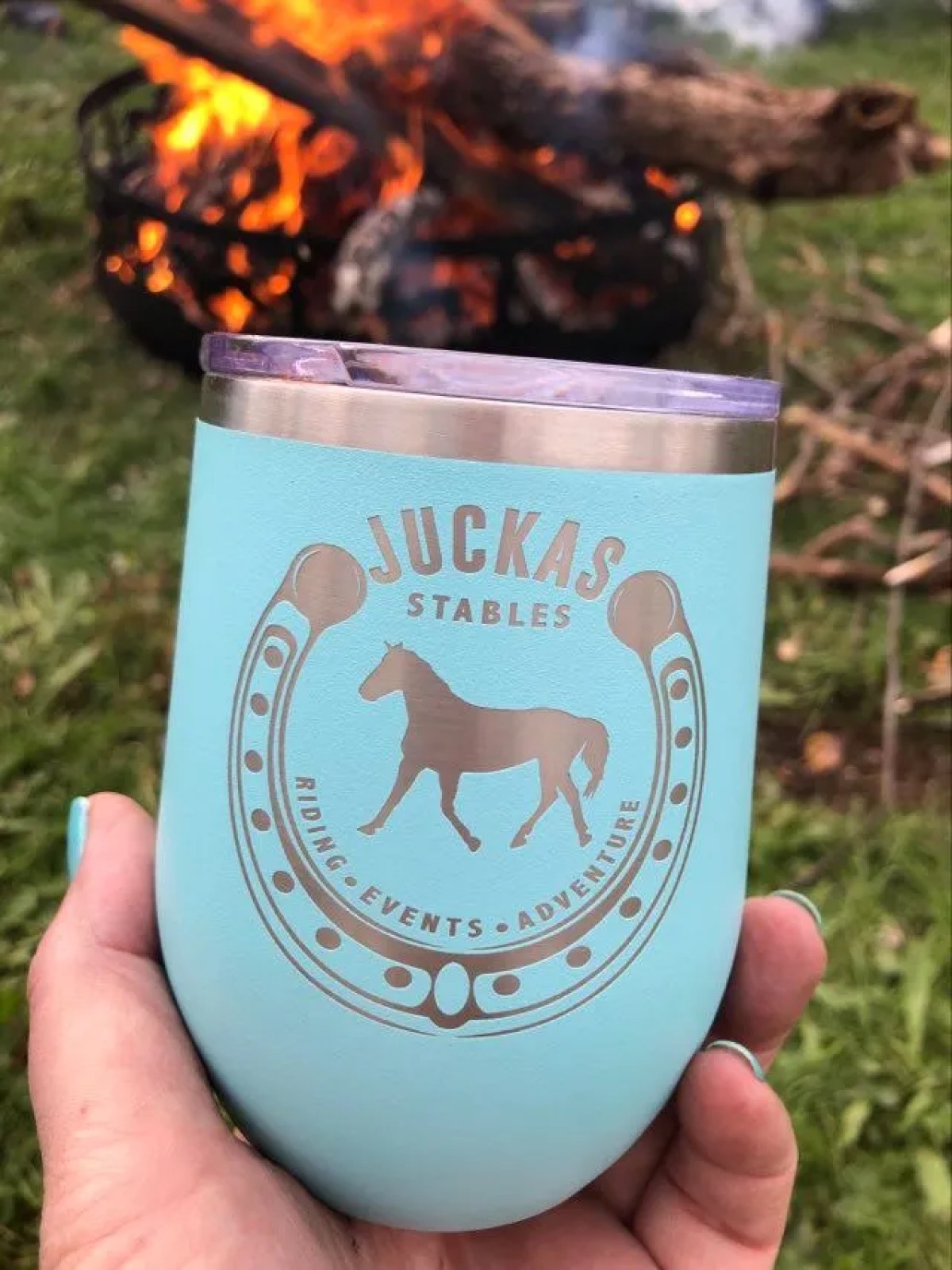 Blue cup with horse logo held near campfire on grass.