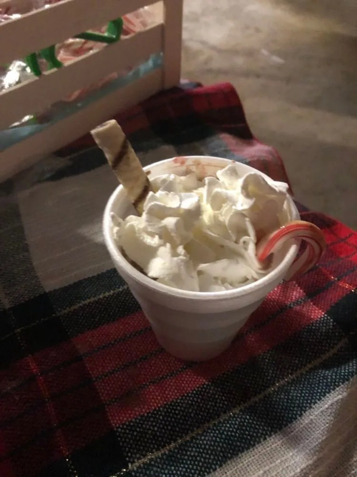 Cup of whipped cream with candy cane and wafer stick on a plaid blanket.