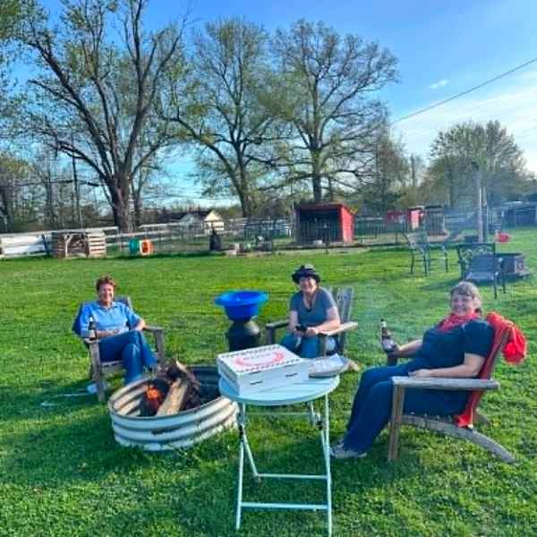 Three people sitting by a fire pit in a yard with trees and a shed.