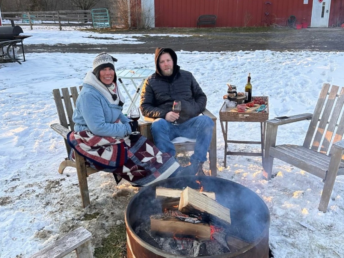 Two people sitting by a fire pit in a snowy setting, holding drinks and wrapped in blankets.